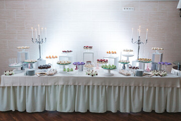 View from distance of white buffet table with delicious colorful desserts. Wedding catering on sweet bar. Patisserie on pre party of luxury gala night