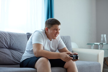 keen caucasian boy playing video games, overweight boy sit on sofa alone, has problems with...