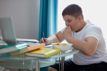 fat overweight boy eat junk food while doing homework, young caucasian boy sit at desk with...