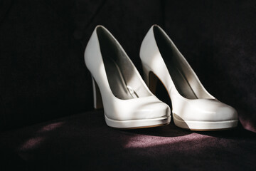 Front view of stylish white wedding shoes on dark background in sunny rays. Fashionable bridal footwear for official occasions, clothes elements. Concept of wedding day preparation, woman clothing.