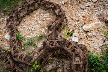 Rusty old chain discarded on stony ground, weeds growing around it. 03295 