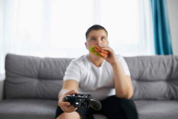 fat caucasian teenager boy enjoys video games and eat junk food, overweight boy sits on sofa...