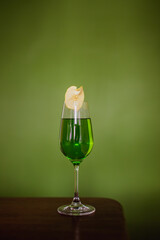 A green cocktail in a flute glass garnished with a dehydrated apple slice on green background