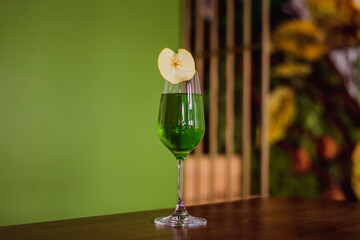 A green cocktail in a flute glass garnished with a dehydrated apple slice on green background