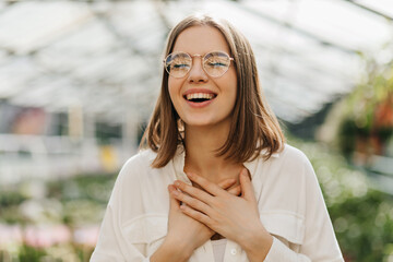 Fototapeta premium Cheerful caucasian girl posing in orangery. Photo of laughing cute woman in glasses having fun in greenhouse.