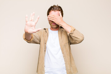blonde adult caucasic man covering face with hand and putting other hand up front to stop camera,...