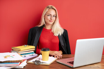 portrait of confident caucasian blonde female sitting at office desk with laptop and papers, young lady in casual wear work as manager, has too much work, think, isolated over red background