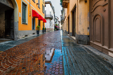 Narrow cobblestone street with puddle among old houses in Alba, Italy.