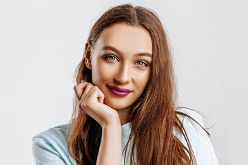 Beautiful sweet brunette girl with long hair with fashion makeup with purple lips on an isolated gray background. The woman holds her hand to her face and looks into the camera. Kind cute model