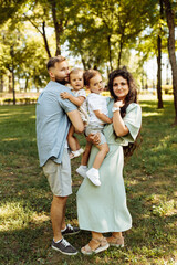 Naklejka na ściany i meble Portrait of lovely family with little kids at the park, caring parents hold beautiful children in arms, smiling, spend time together, enjoy happy moments, parenting concept
