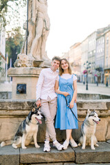 Naklejka na ściany i meble Happy Caucasian young couple in love, with two husky dogs, posing to camera in the center of ancient European city standing near old fountain on stone stairs. Couple in love with dogs in the city