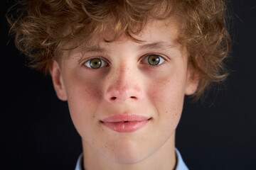 portrait of shy freckled curly boy looking at camera, open-minded boy in studio with black...