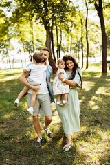 Naklejka na ściany i meble Loving parents with little kids walking at the park, adorable mother hold in arms beautiful daughter, cute son sitting in fathers hands, enjoy happy family weekend concept
