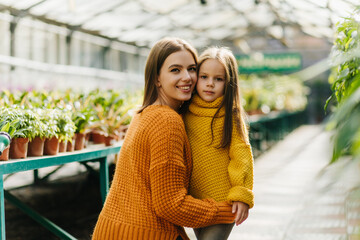 Magnificent european woman embracing daughter in knitted sweater. Young mother and child posing near plants in pots.