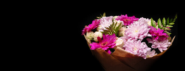 Large beautiful bouquet of chrysanthemums, gerberas, roses and ferns in pink and purple colors, packed in brown craft paper and isolated on a black background. Postcard for the holiday.
