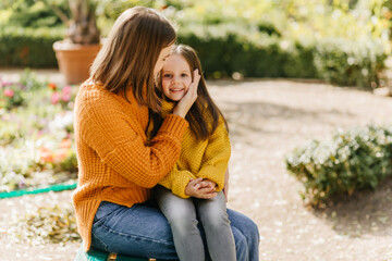 Trendy woman kissing child with love. Lovely kid in yellow sweater sitting on mother's knees in park.