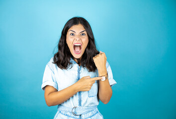 Young beautiful woman wearing a denim jumpsuit over isolated blue background surprised and pointing...