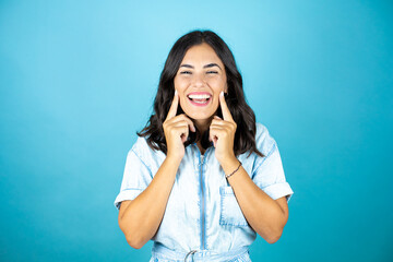 Young beautiful woman wearing a denim jumpsuit over isolated blue background smiling confident showing and pointing with fingers teeth and mouth