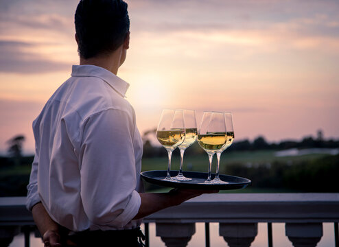 Waiter in white shirt holding tray of four wine glasses looking at sunset and golf course