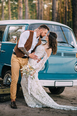 Boho-style newlyweds hug tenderly against the background of a blue vintage minibus. Full-length...