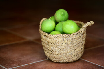 There are green organic lemons in a small woven basket against a dark background on brown tiles