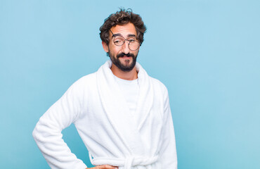 young bearded man wearing a bath robe smiling happily with a hand on hip and confident, positive,...