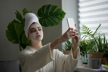 Young woman with cream mud mask on her face taking selfie on her phone.