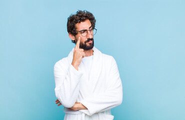 young bearded man wearing a bath robe with a concentrated look, wondering with a doubtful...