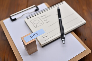 A note with the user's persona written on it is placed on a clipboard along with a pen. A sticky note stamped with "HCD" is placed on a wooden cube.