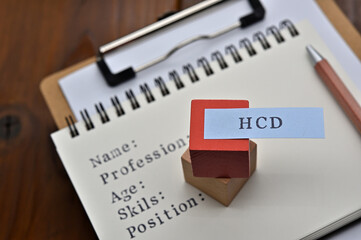 A note with the user's persona written on it is placed on a clipboard along with a pen. A sticky note stamped with "HCD" is placed on a wooden cube.