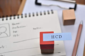 A note with the user's persona written on it is placed on a clipboard along with a pen. A sticky note stamped with "HCD" is placed on a wooden cube.
