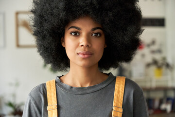 Confident African American gen z hipster female student with Afro hair looking at camera standing indoors, at home, in modern creative office. Mixed race young woman close up headshot portrait.
