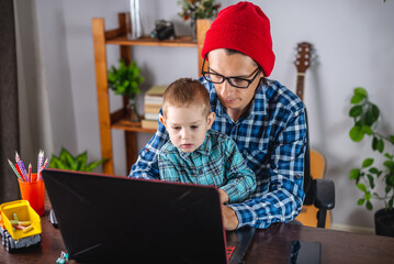 Modern man is working on a laptop, and his little son is sitting on his lap. Concept of family and remote work from home