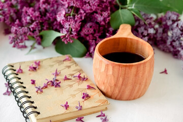 Fototapeta na wymiar Wooden finnish cup with coffee on the table. Nearby lies a closed notebook for notes and a bouquet of lilacs. Good morning and good mood, planning a day at breakfast, concept.