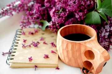 Obraz na płótnie Canvas Wooden finnish cup with coffee on the table. Nearby lies a closed notebook for notes and a bouquet of lilacs. Good morning and good mood, planning a day at breakfast, concept.