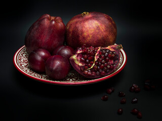 Still life in dark red tints with pomegranate, plum and apple on a Uzbeq-style plate with dark background