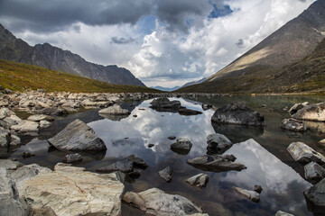 A cloudy sky is reflected in a shallow mountain lake with many stones along the shores. The...