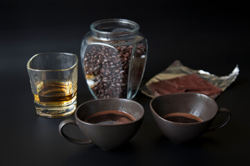 Two dark cups of coffee on a black background. Glass jar with coffee beans, a glass of whiskey, and a chocolate bar are out of focus in the background.