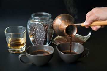 Two dark cups of coffee on a black background. A hand pours coffee from a Turkish coffee pot. Glass jar with coffee beans, a glass of whiskey, and a chocolate bar are out of focus in the background.