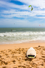 Obraz premium Surfboard on a sand beach. Kitesurfers in the sea in the background. Blue sky with light clouds