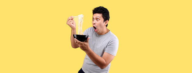  Close up Asian man eating yummy hot and spicy instant noodle using chopsticks and bowl isolated on yellow background in studio With copy space.