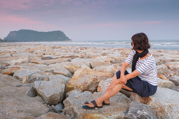 An Asian woman in a T-shirt and sunglasses is relaxing by the sea on a cold evening.