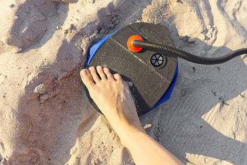A woman with air foot pump pumps an inflatable mattress or air bed at sandy beach. Foot inflates...
