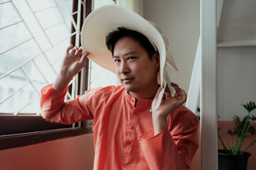 Gay in orange mandarin collar try the white hat with ribbon while he sit at the window.