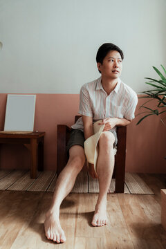Asian gay sitting on the wooden chair with white hat looking outside the window.