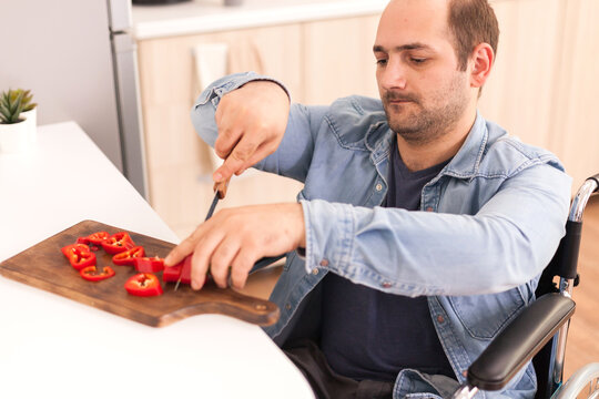 Man with mobility handicap slicing red pepper using sharp knife in kitchen. Disabled paralyzed handicapped man with walking disability integrating after an accident.