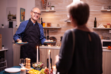 Senior man smiling at wife while holding plates with food food their romantic relationship anniversary. Elderly old couple talking, sitting at the table in kitchen, enjoying the meal,
