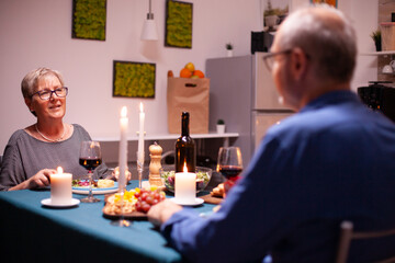 Mature woman having a conversation with husband while holding glass of red wine in kitchen. Senior couple sitting at the table in kitchen, talking, enjoying the meal, celebrating their anniversary in