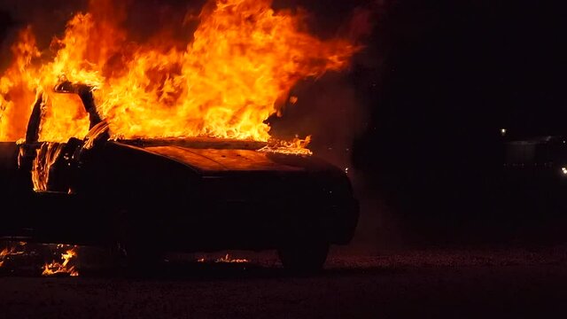 burning car at night, right side, half diagonal from the front, fire department approaching in the background