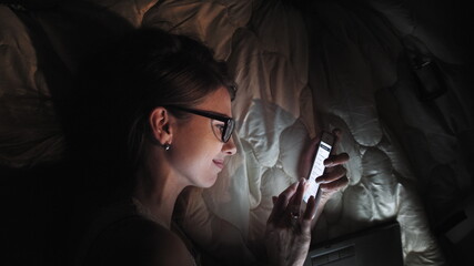 Teenage Girl on Touch Screen Smart Laptop and Headphones on Bed, Student in Dark Room with Mobile Night Light, a Young Woman studying and reading in the evening.